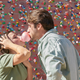 Katie and Greg blowing bubbles with gum in front of a big wall covered in gum